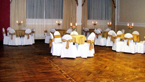 A banquet hall with rows of tables and chairs, each with a white tablecloth and a gold sashed chair. - Coloso Potosi (Potosi)