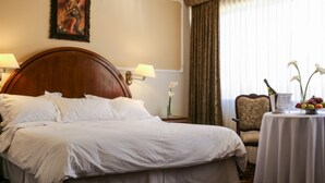 A hotel room with a large bed, two bedside lamps, a chair, a small table with fruit and flowers, and a window with curtains. - Coloso Potosi (Potosi)