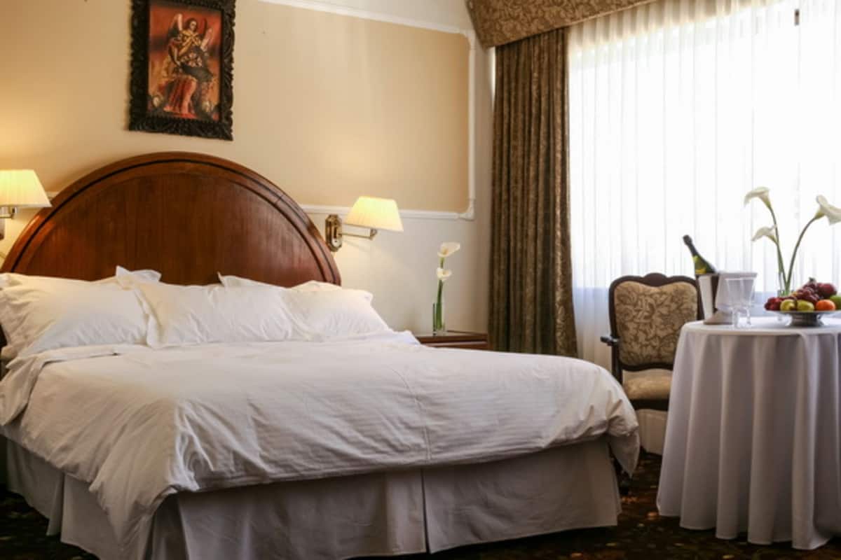 Habitación de hotel con una cama grande, dos lámparas de noche, una silla, una mesita con fruta y flores, y una ventana con cortinas.