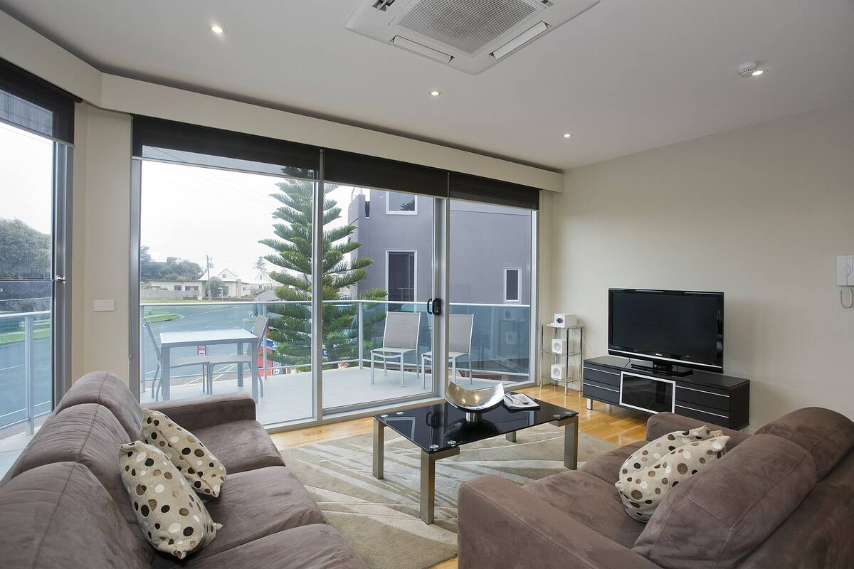 A modern living room with a sofa set, a TV, and a glass door leading to an outdoor patio with a table and chairs.