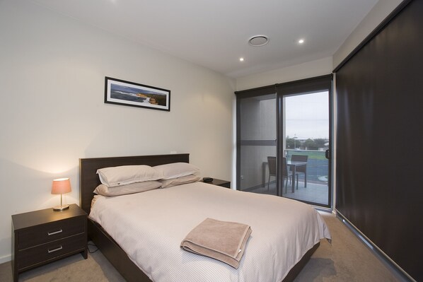 A bedroom with a bed, a nightstand with a lamp, a framed picture on the wall, and a sliding glass door leading to a balcony.