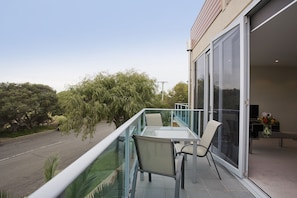 A balcony with a glass table and chairs, a sliding glass door, and a view of trees and a road. - 7 Bell Street Apartments (Torquay)