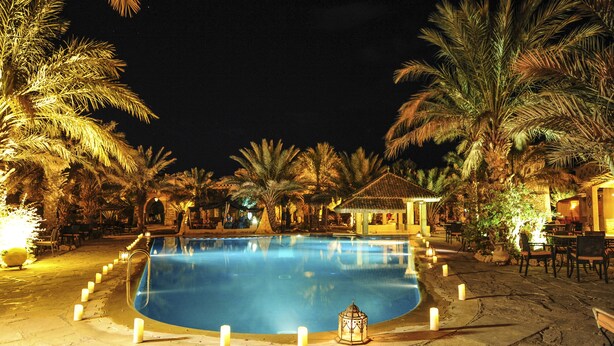 A nighttime swimming pool surrounded by palm trees and outdoor seating areas. - Kasbah Hotel Xaluca Arfoud (Aarab Sebbah Ziz)