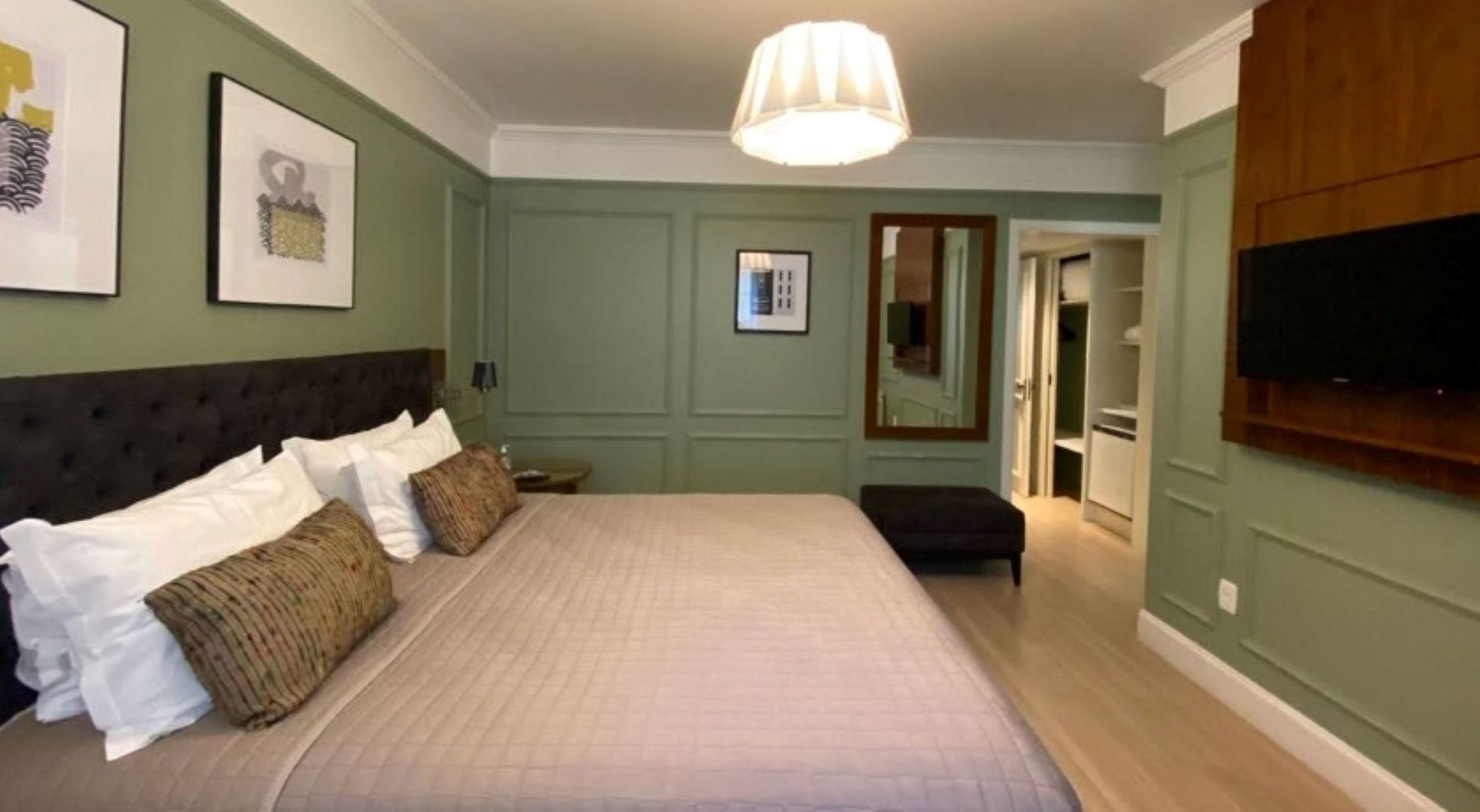 Um quarto com uma cama grande, uma mesinha de cabeceira, um espelho, uma TV fixada na parede e uma porta que dá para outro cômodo.