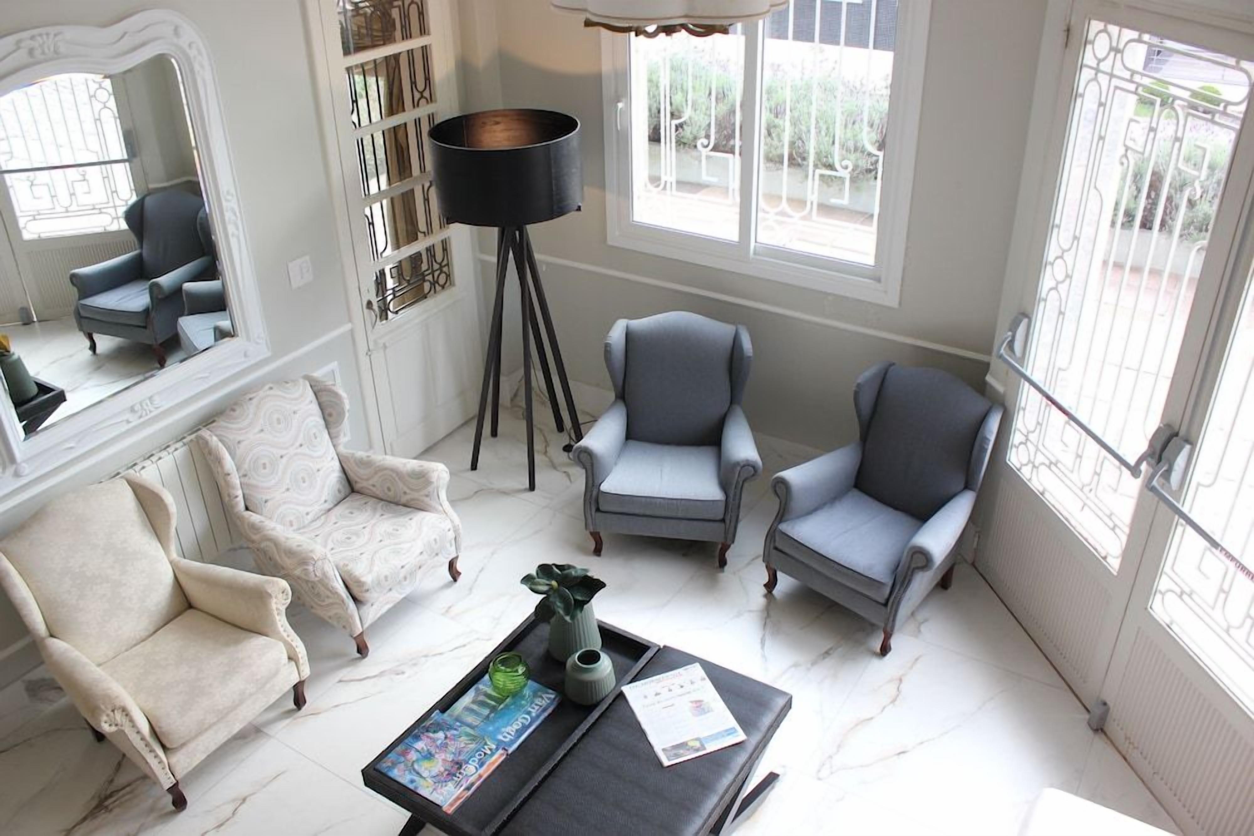 Uma sala de estar com um sofá branco, duas poltronas, uma mesa de centro com revistas e um abajur.
