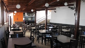 A spacious restaurant with multiple tables and chairs, wooden ceiling beams, and hanging light fixtures.
