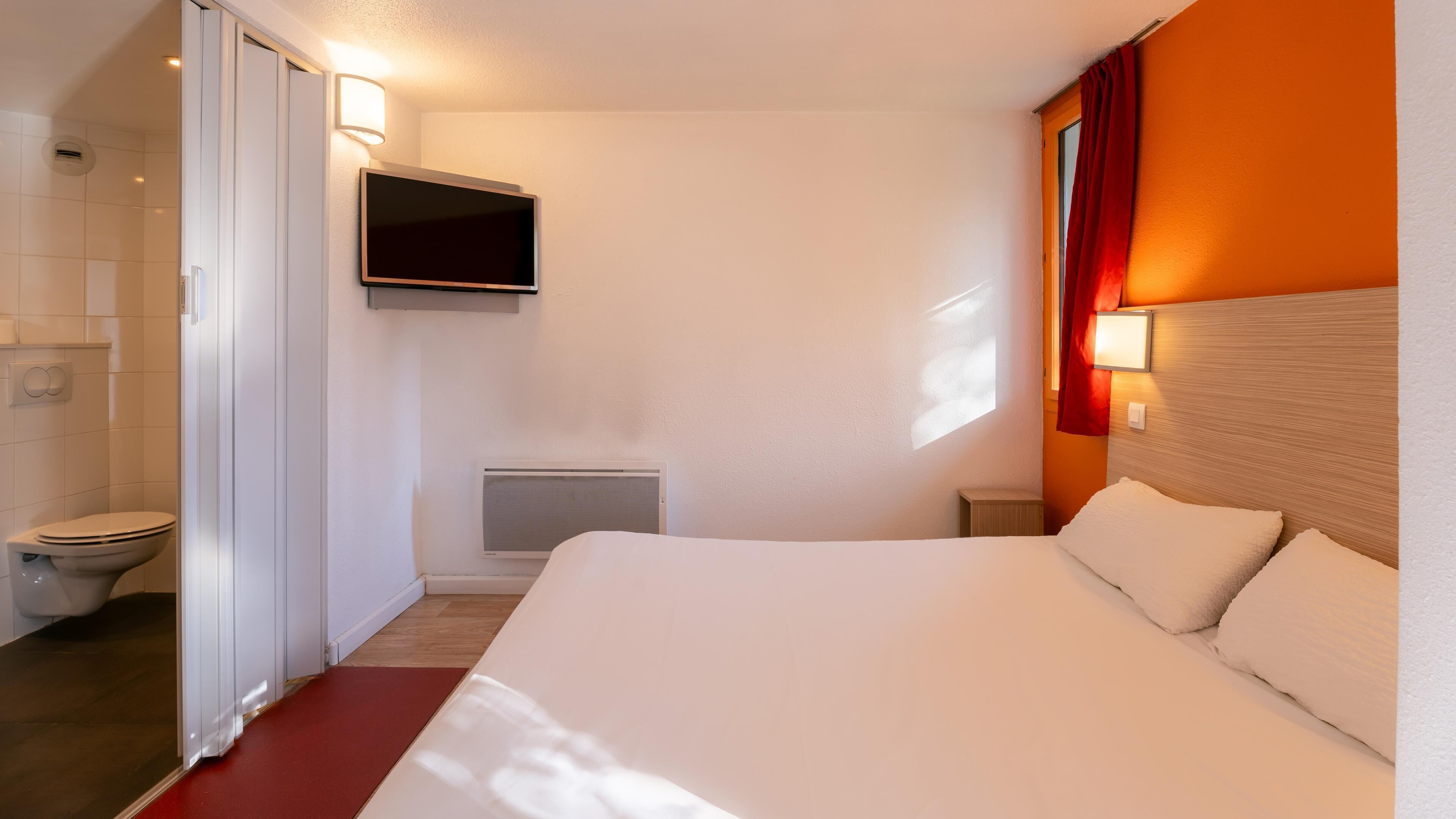 Una habitación de hotel con una cama, televisión en la pared, radiador y ventana con cortinas.