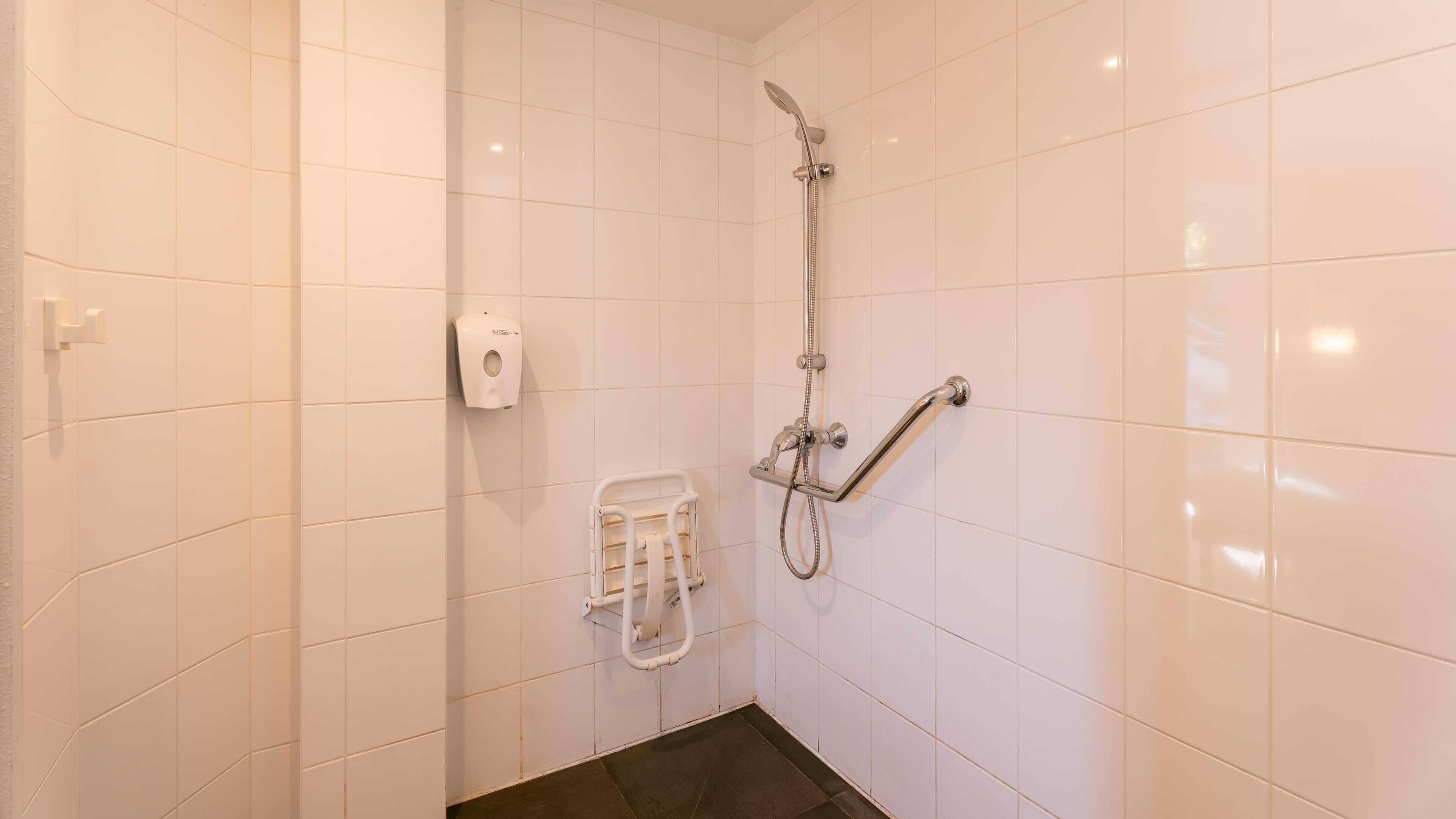 Un baño pequeño con azulejos blancos, ducha, dispensador de jabón y barra de toallas.