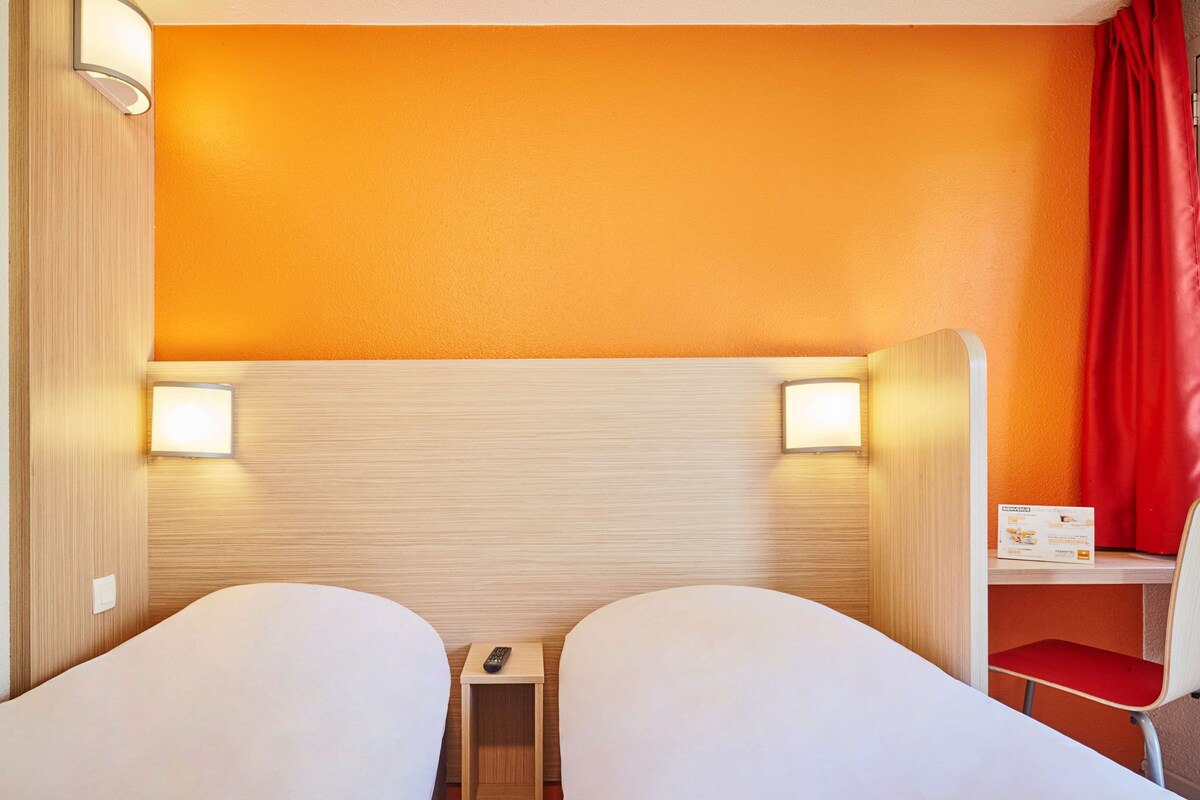 A hotel room with two beds, a small table, a chair, and a red curtain.