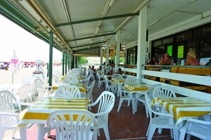 An outdoor restaurant with white plastic chairs and yellow-striped tables, covered by a roof with green beams. - Village Camping Joker (Cavallino-Treporti)