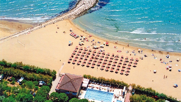 Une vue aérienne d’une plage avec des rangées de chaises longues et de parasols, une piscine et un bâtiment doté d’un toit en pente.