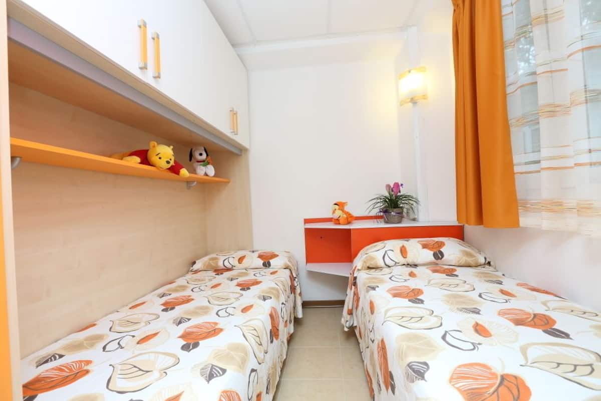 Une petite chambre agréable, dotée de deux lits simples, d’un bureau avec une chaise et d’une étagère où sont posés des peluches.