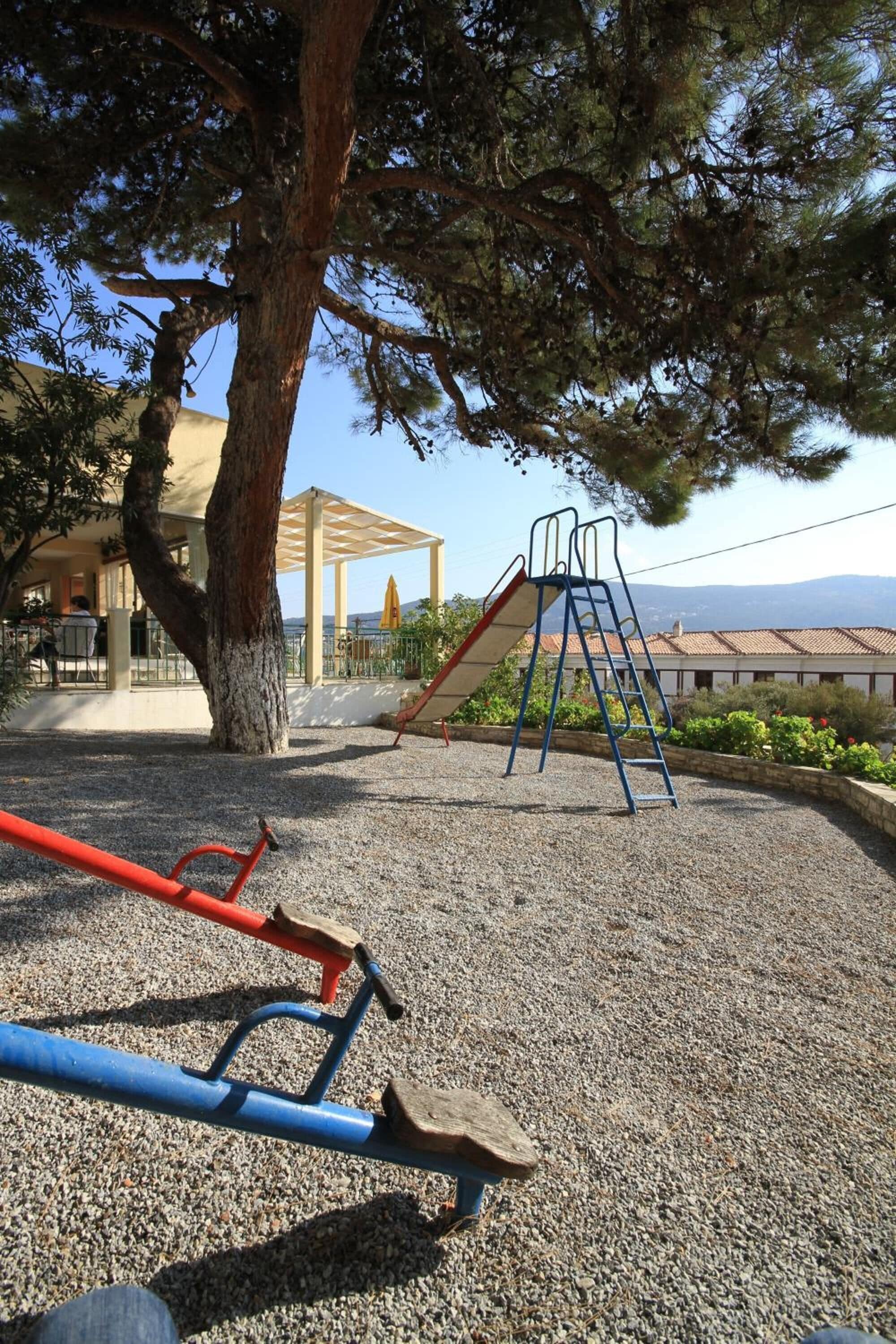 Un parque infantil con un tobogán y una estructura para escalar, rodeado de árboles y un edificio con un área cubierta.