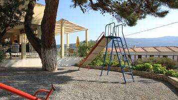Un parque infantil con un tobogán y una estructura para escalar, rodeado de árboles y un edificio con un área cubierta.
