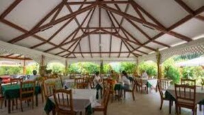 Un comedor interior con mesas y sillas de madera, un techo alto con vigas de madera y vistas al exterior con vegetación.