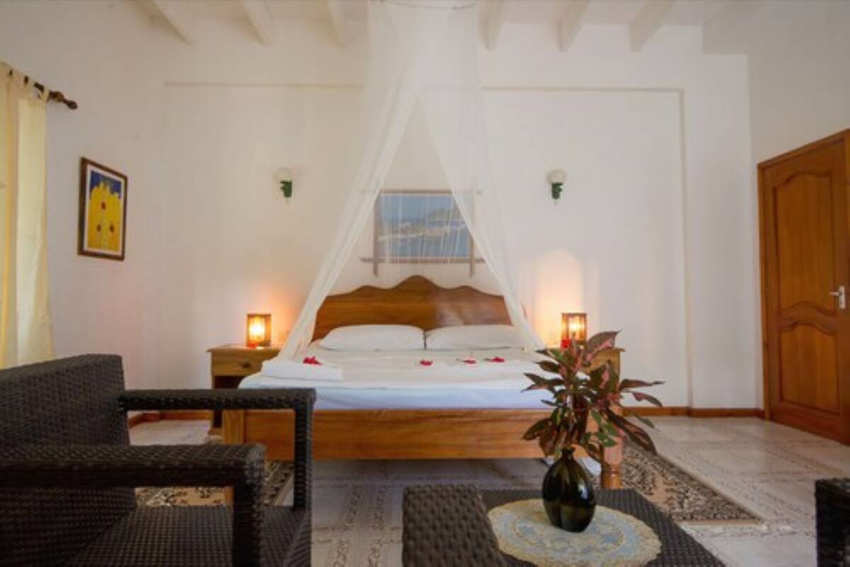 Un dormitorio con cama de madera, ropa de cama blanca, dosel, dos lámparas de noche, planta en maceta y una silla de mimbre.