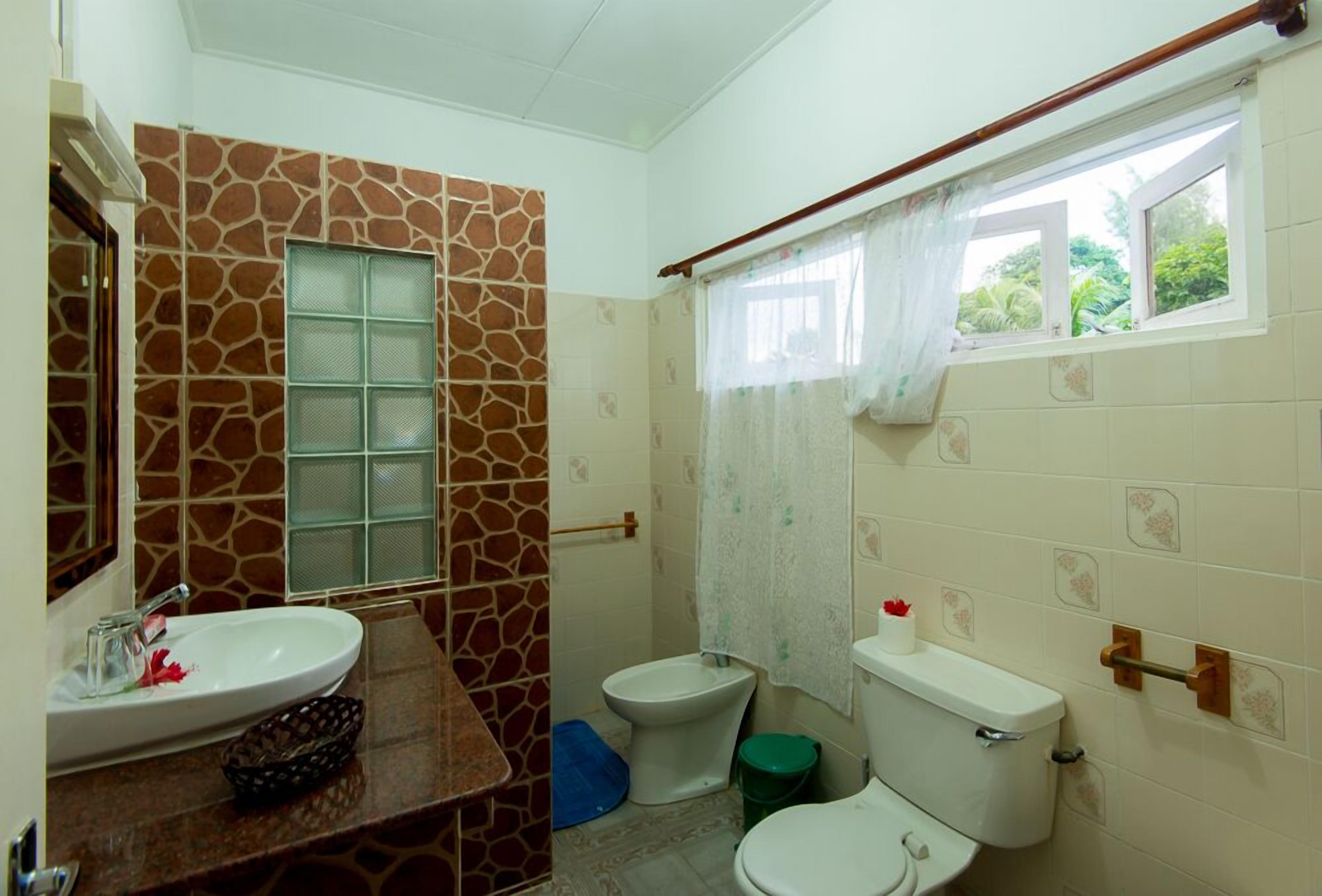 Ein Badezimmer mit einer Wand im Steinoptik, einem Waschbecken, einem Bidet, einer Toilette und einem Fenster mit Vorhang.