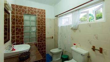 Ein Badezimmer mit einer Wand im Steinoptik, einem Waschbecken, einem Bidet, einer Toilette und einem Fenster mit Vorhang.