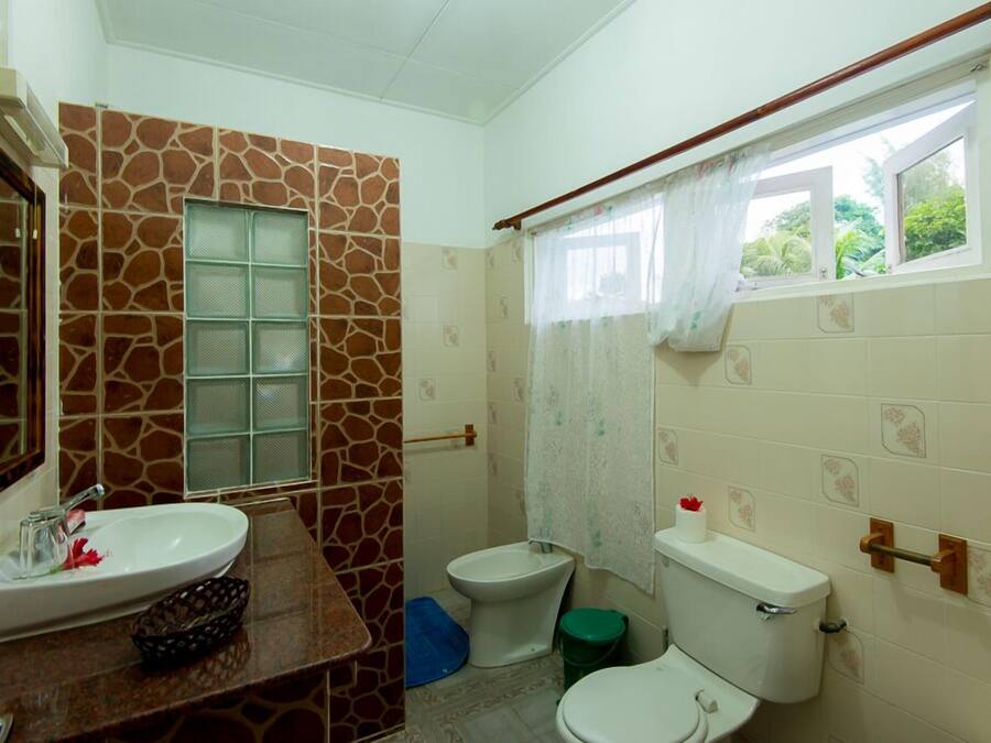 Ein Badezimmer mit einer Wand im Steinoptik, einem Waschbecken, einem Bidet, einer Toilette und einem Fenster mit Vorhang.