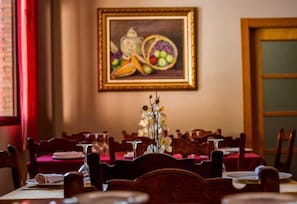 A dining area with wooden chairs and tables, a vase with flowers, and a painting of fruit on the wall. - El Nacimiento (Deifontes)