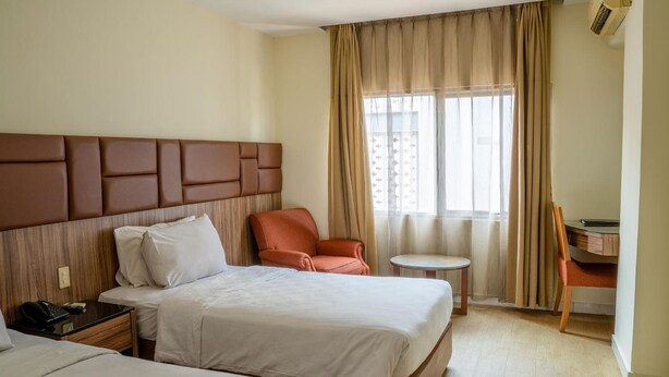 A hotel room with two beds, a wooden headboard, a small table, a chair, and a window with curtains. - Alia Cikini (Jakarta)
