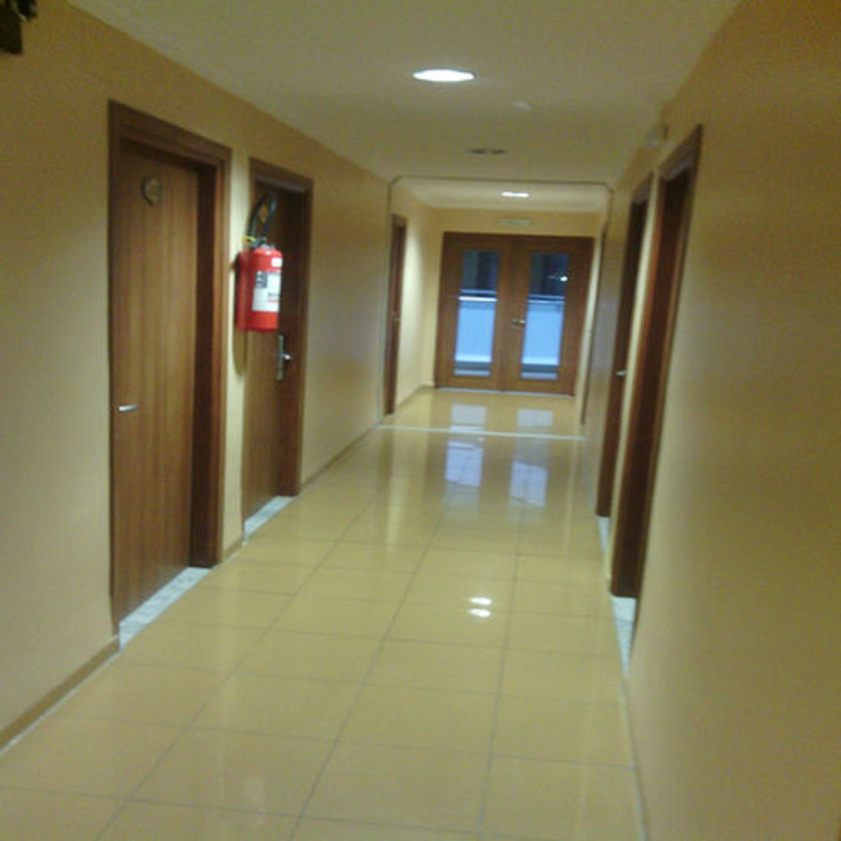 A hallway with wooden doors, a fire extinguisher mounted on the wall, and a tiled floor.