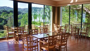 Un comedor con varias mesas y sillas, ventanas amplias con vistas a una ladera boscosa.