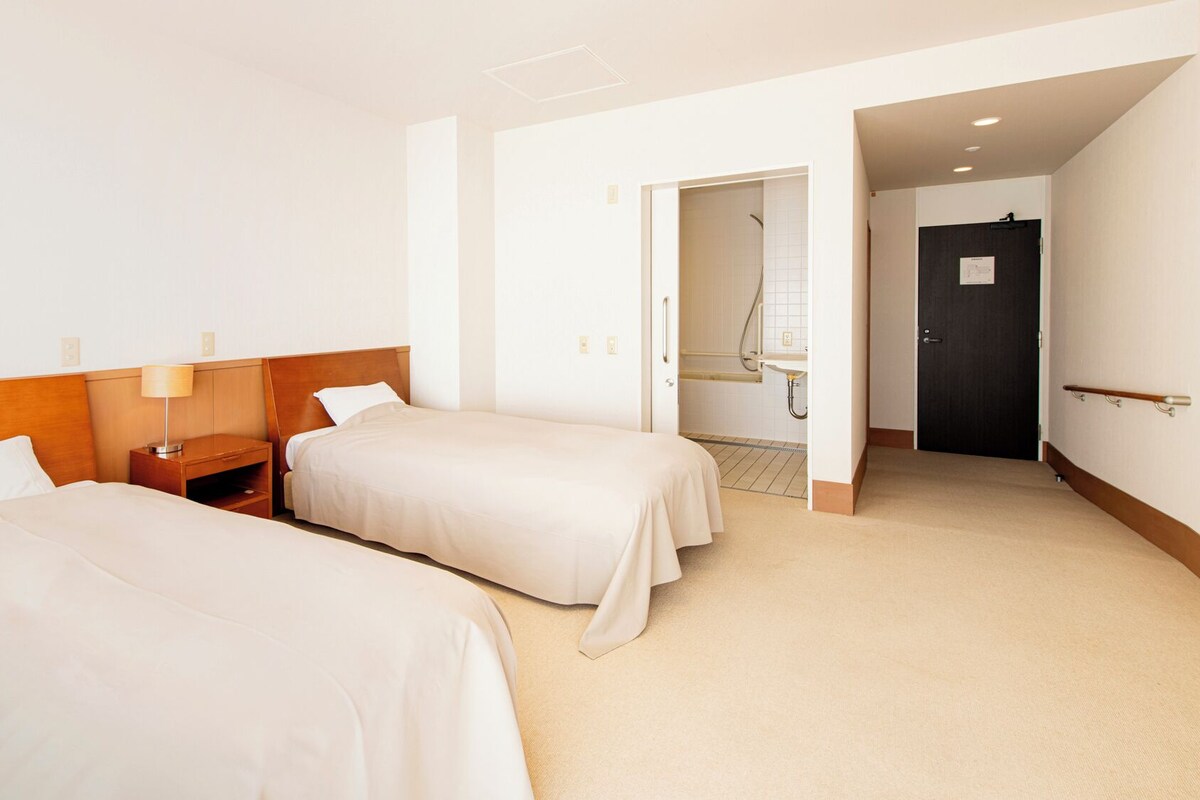 Habitación de hotel con dos camas, una mesita de noche, una lámpara y un baño visible a través de una puerta abierta.