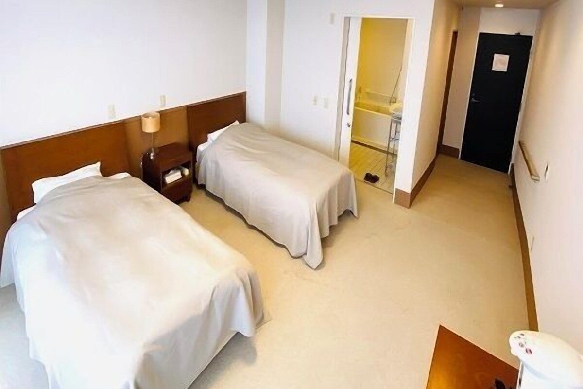 Habitación de hotel con dos camas, una mesita de noche, una lámpara, un baño con ducha y una puerta que da a otra habitación.