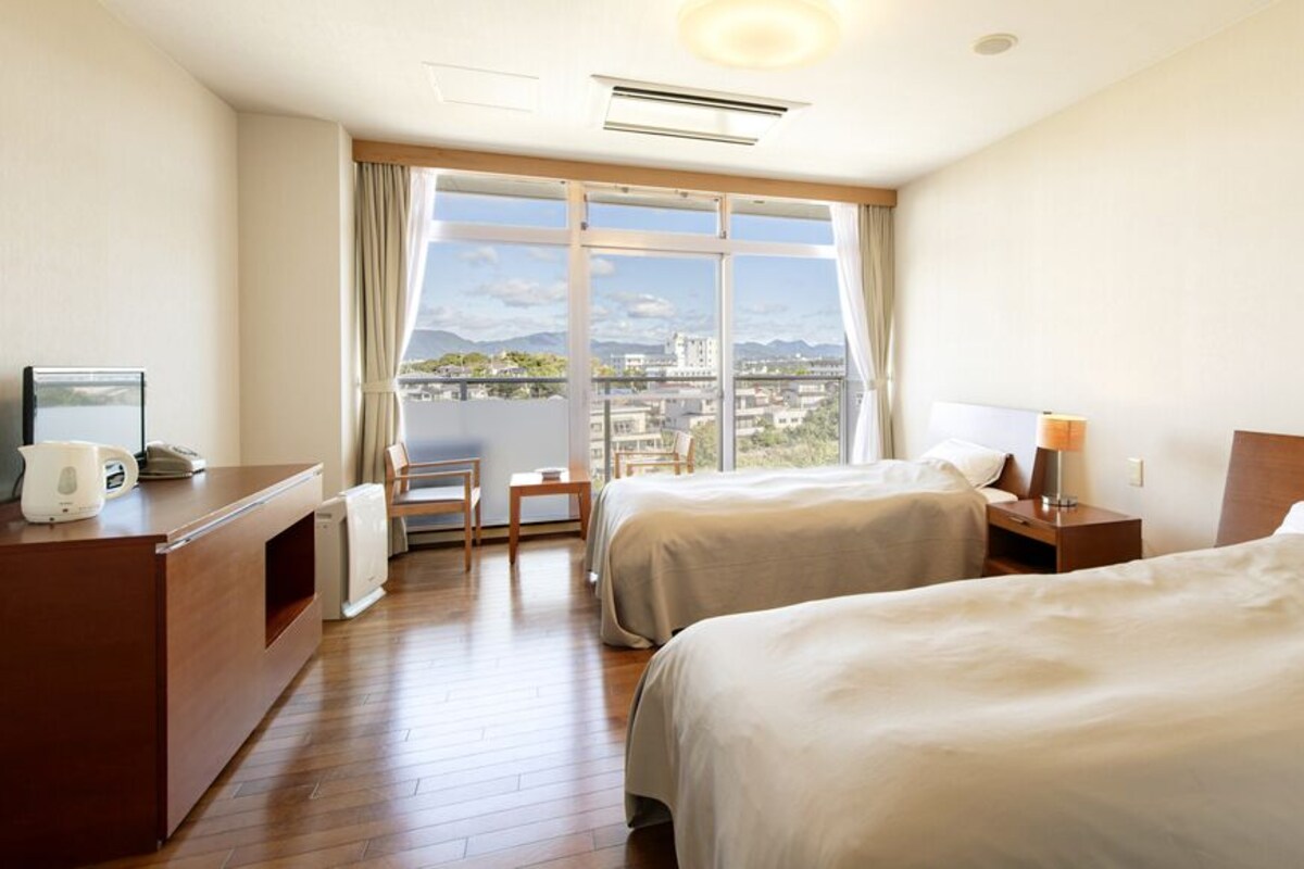 Habitación de hotel con dos camas, un escritorio, una silla, una ventana con vista a la ciudad y un balcón.