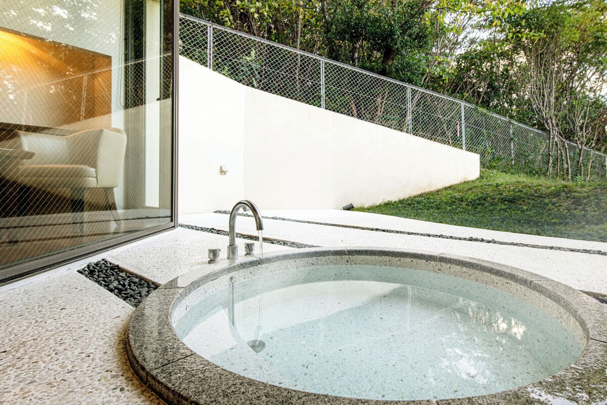 Un baño al aire libre moderno con un jacuzzi circular, zona de ducha con cerramiento de vidrio y valla metálica.