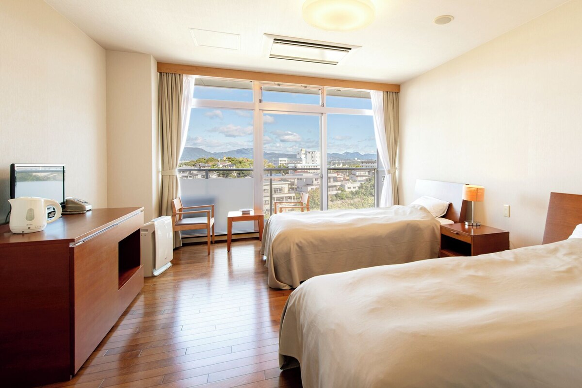 Habitación de hotel con dos camas, un escritorio, una silla, una mesa, una lámpara y vistas a la ciudad.