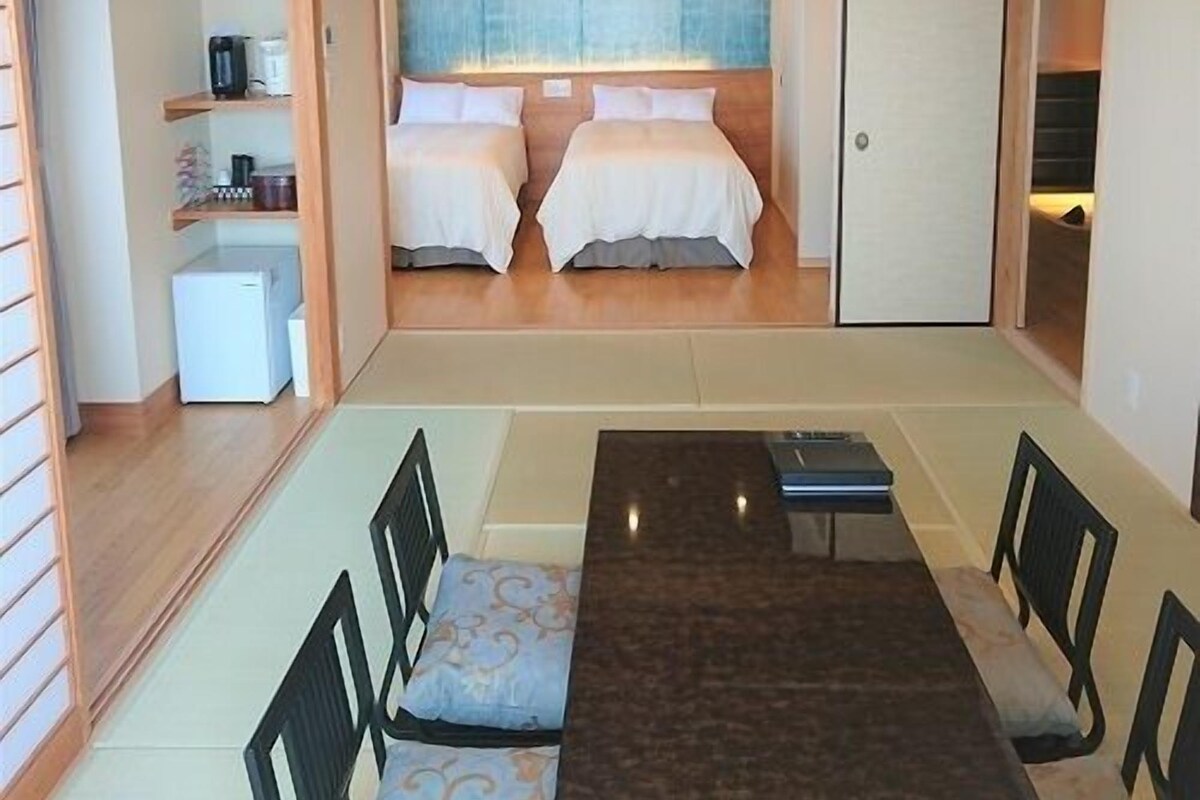 Habitación compacta con dos camas, una kitchenette pequeña y un espacio de descanso con sillas y una mesa.