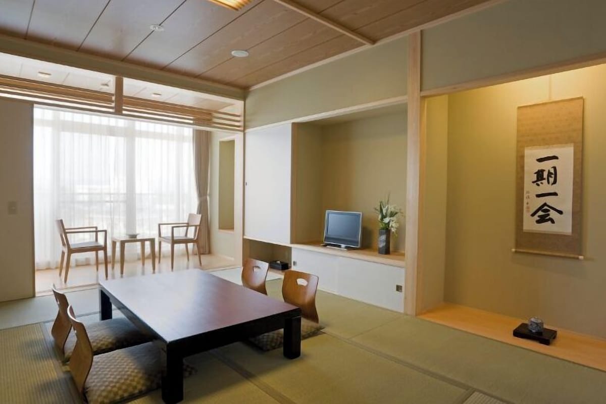 Una habitación tradicional japonesa con suelo de tatami, una mesa baja, sillas, un televisor y una obra de caligrafía en la pared.