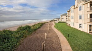 Un paseo marítimo con vista a la playa, un área con césped y una hilera de edificios de varios pisos color beige con balcones.