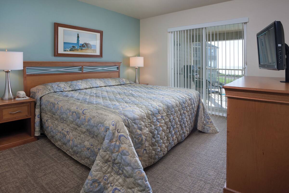 Quarto de hotel com uma cama grande, duas mesas de cabeceira com abajures, uma televisão em uma cômoda de madeira, um quadro na parede e uma porta de vidro corrediça para uma varanda.