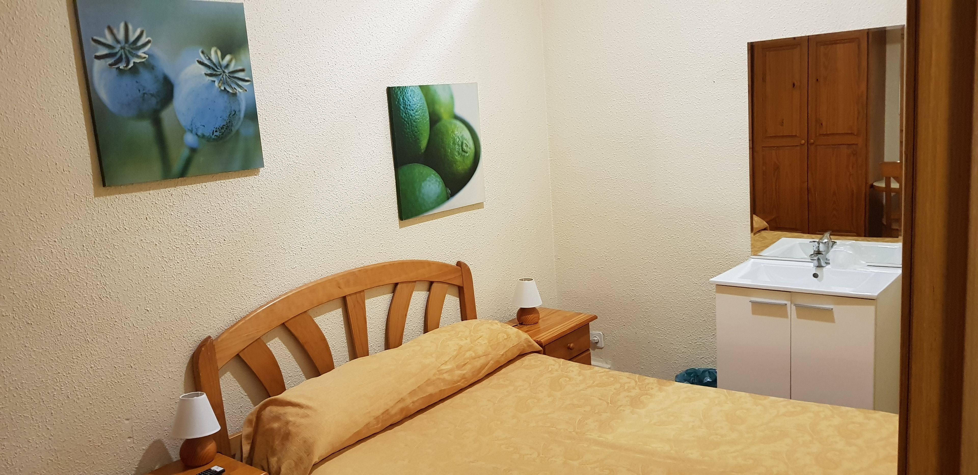 Un dormitorio con cabecera de madera, una cama con manta color beige, una mesita de noche con lámpara, un lavamanos empotrado en la pared y armarios de madera.