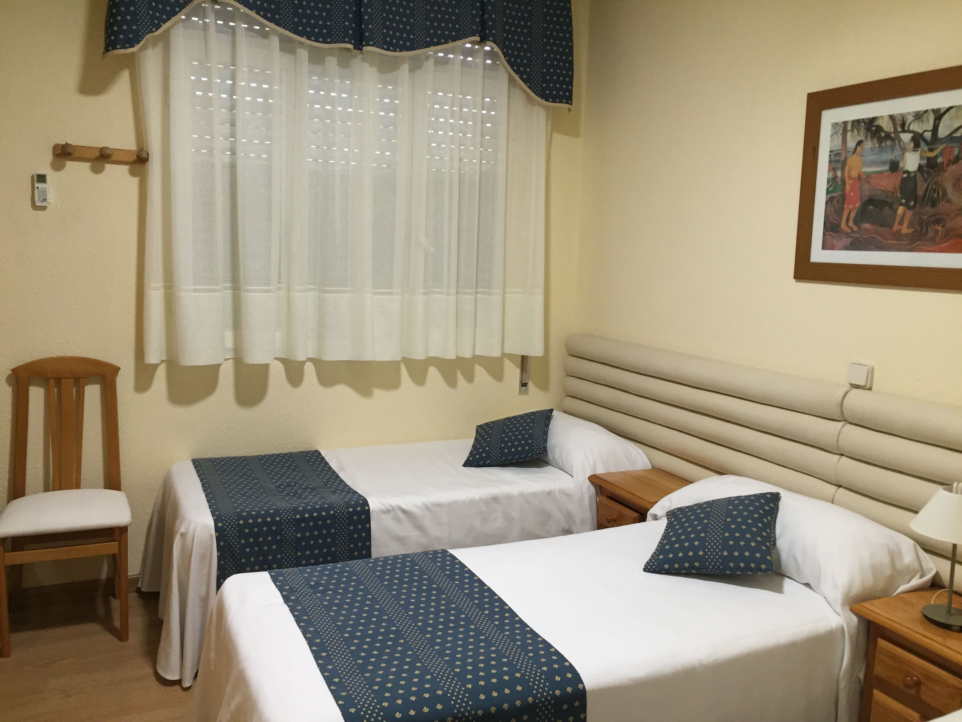 Habitación de hotel con dos camas, una silla de madera, una mesita de noche y un cuadro en la pared.