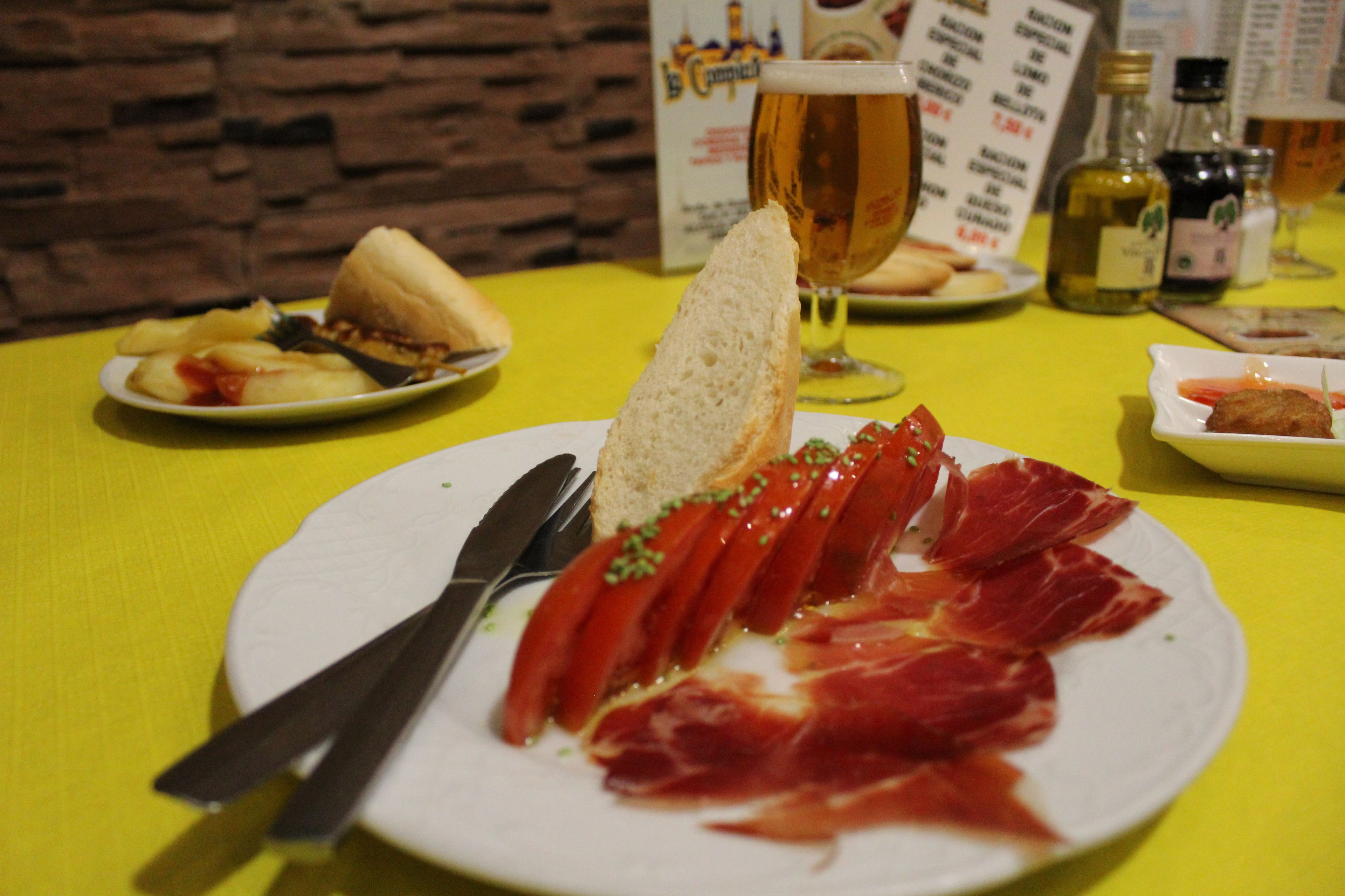 A plate of sliced meat with a knife and fork, a glass of beer, and a sandwich on a yellow tablecloth.