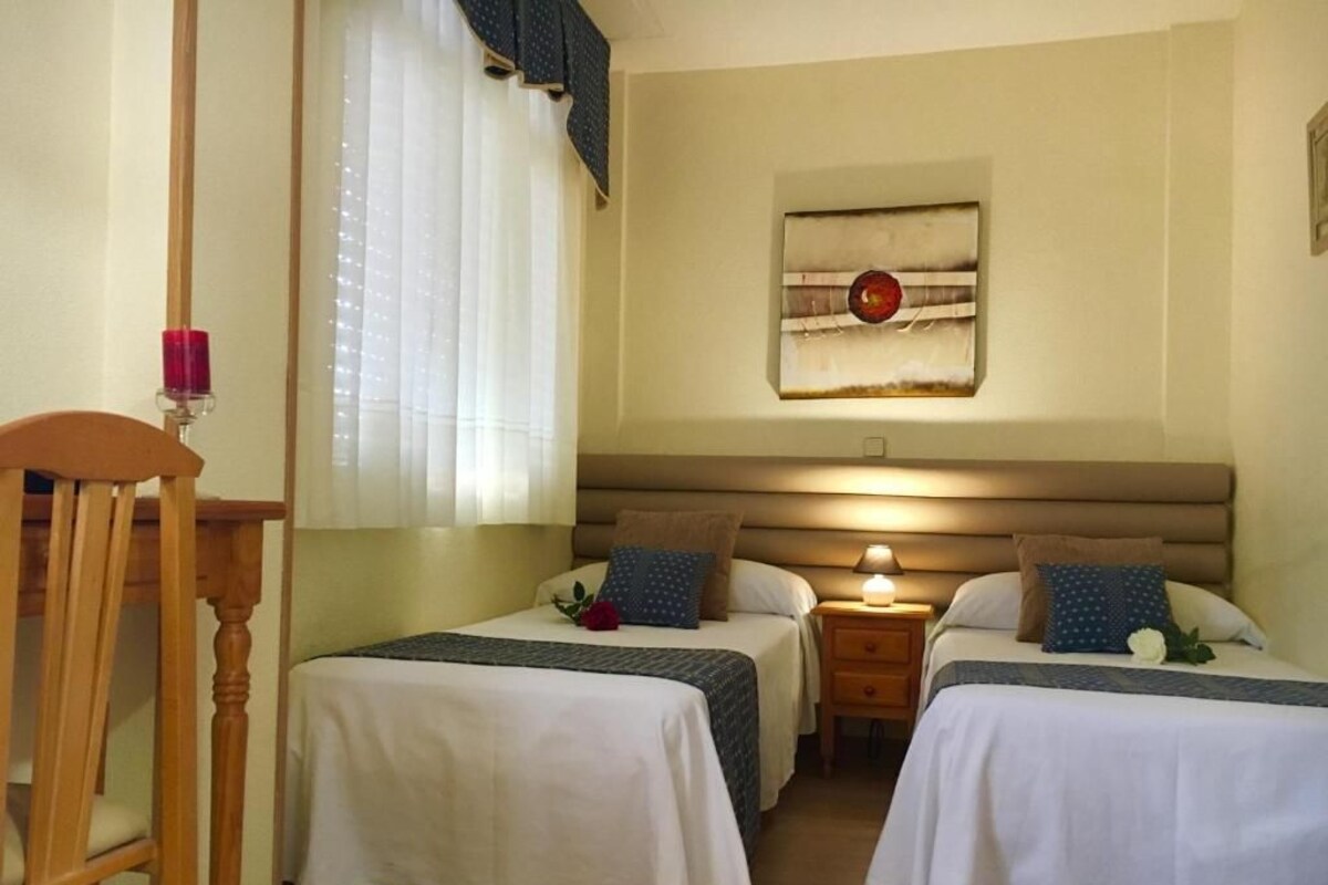 A hotel room with two beds, a wooden chair, a bedside table with a lamp, and a window with curtains.