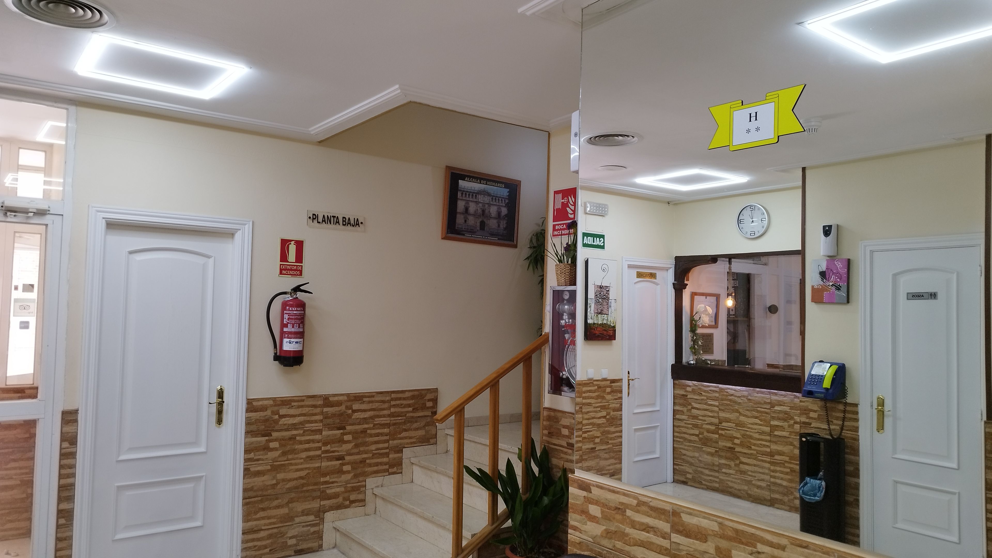 A hotel lobby with a staircase, a fire extinguisher, a clock, and a sign indicating the location.