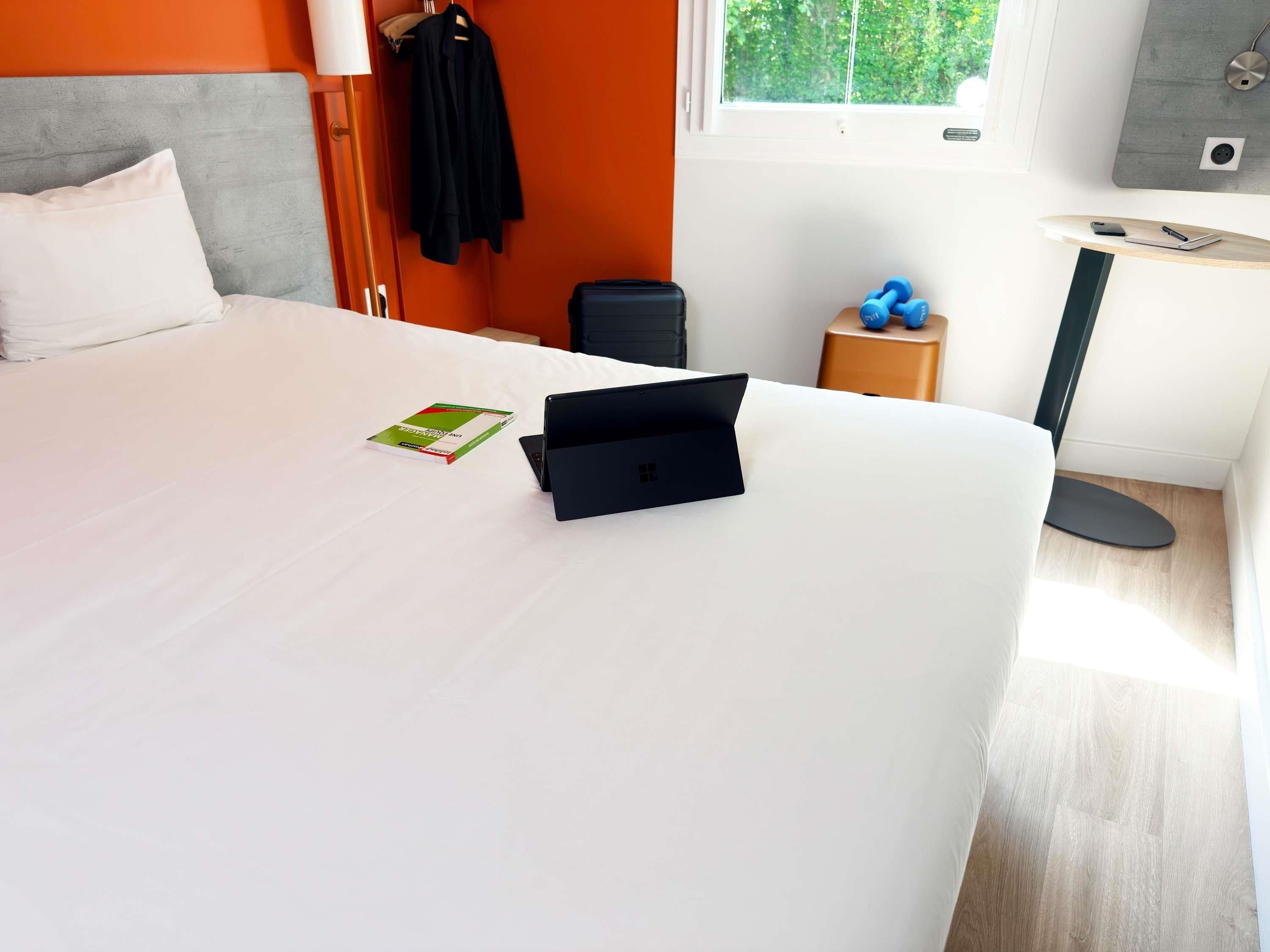 Une chambre d’hôtel avec un lit, un ordinateur portable, un livre, une petite table avec une lampe et une fenêtre donnant sur un paysage verdoyant.