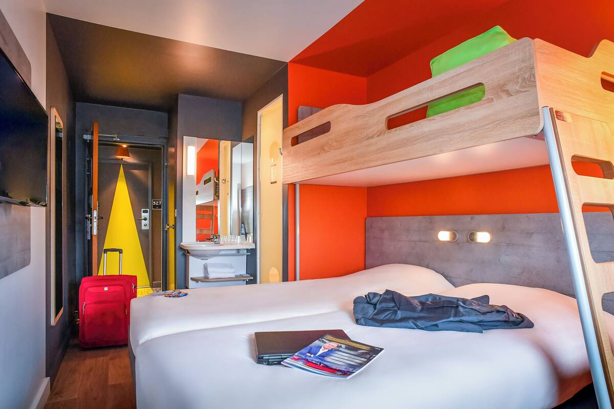 A hotel room with a bunk bed, a single bed, a suitcase, and a magazine on the bed.
