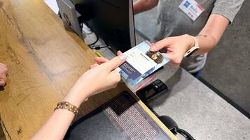 Une personne tenant une carte à un comptoir, devant un écran d’ordinateur et un téléphone.