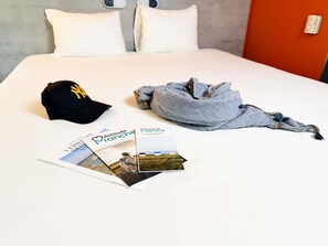 A bed with white linens, a black cap, a grey scarf, and two travel brochures. - Ibis Budget Coutances (Coutances)