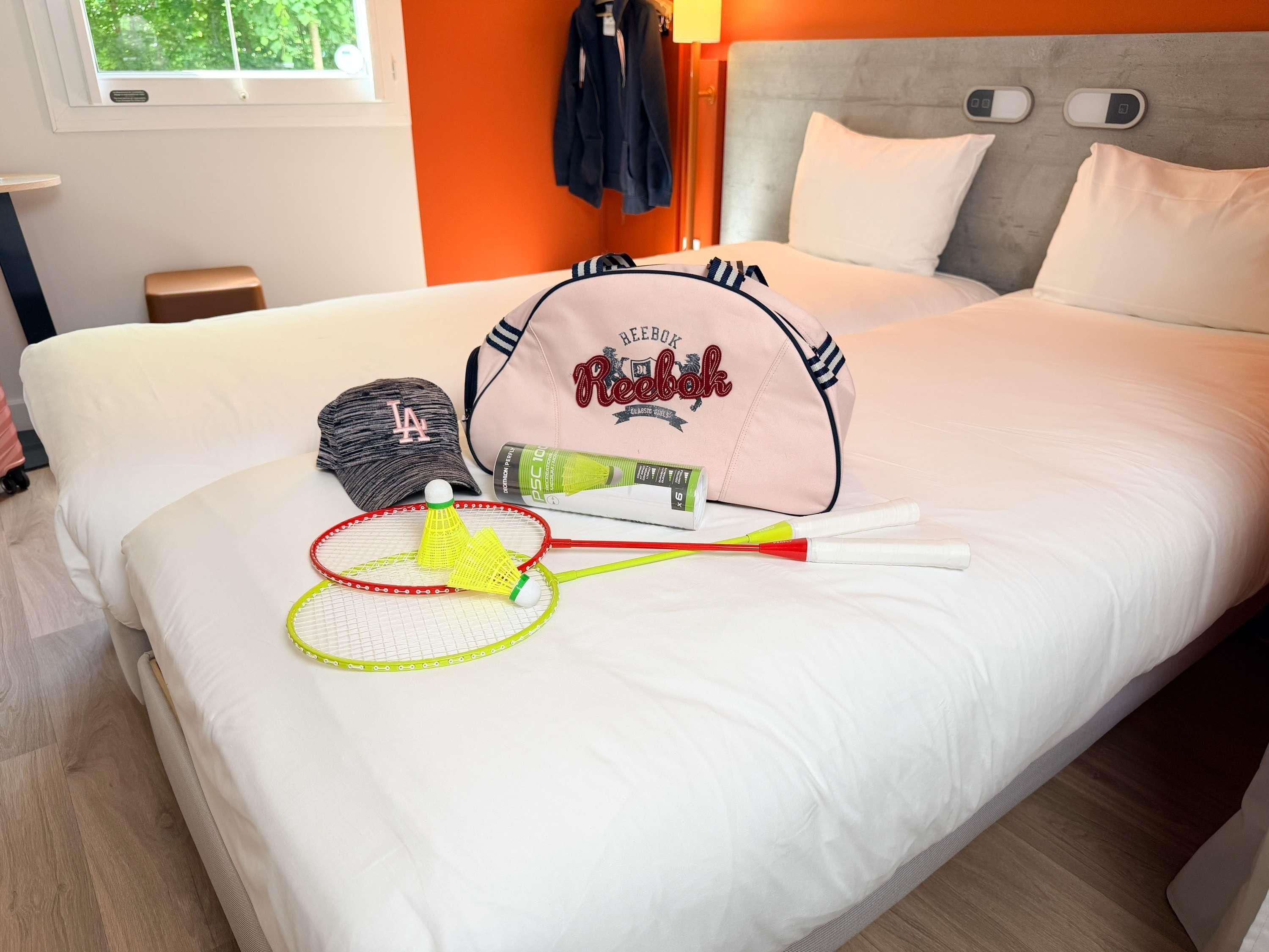 A neatly made bed with a white comforter, a Reebok-branded bag, a cap, a shuttlecock, and a badminton racket.