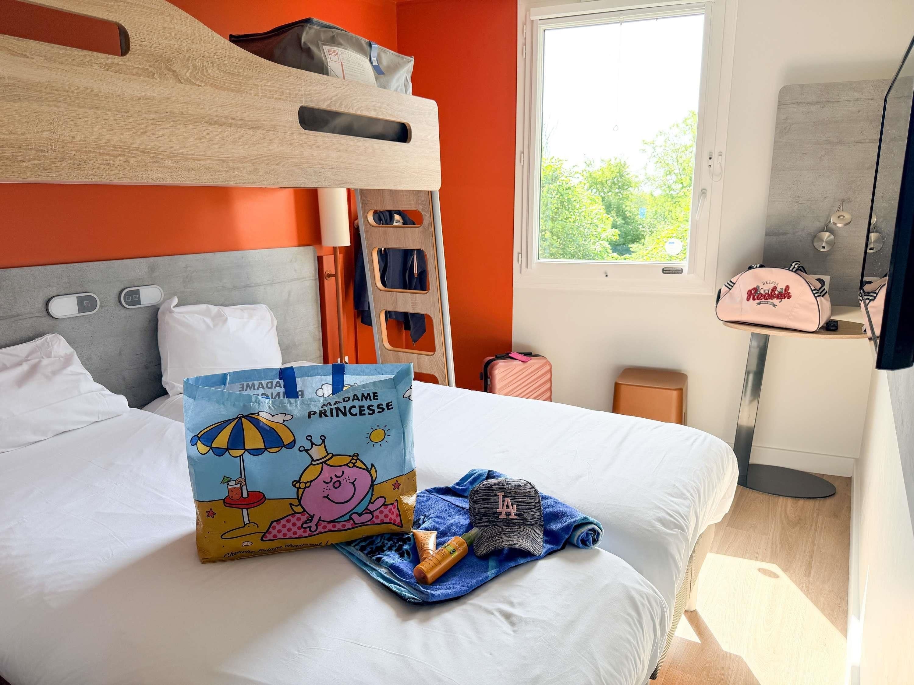 A hotel room with a bunk bed, a window with a view of trees, a bedside table with a Reebok bag, and a bag with a cartoon character on it.