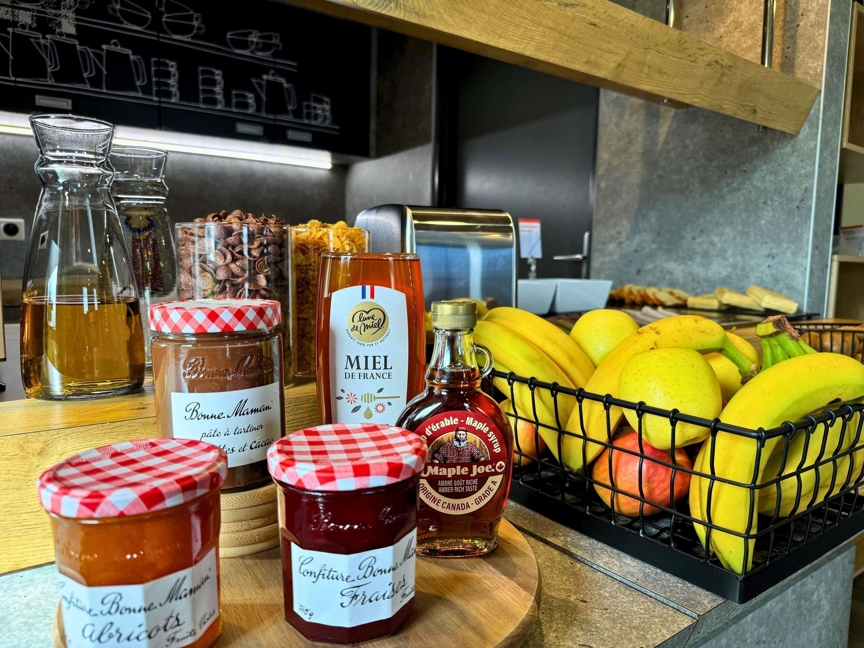 A kitchen counter with various food items including jars of preserves, a bottle of maple syrup, and a basket of bananas and apples.