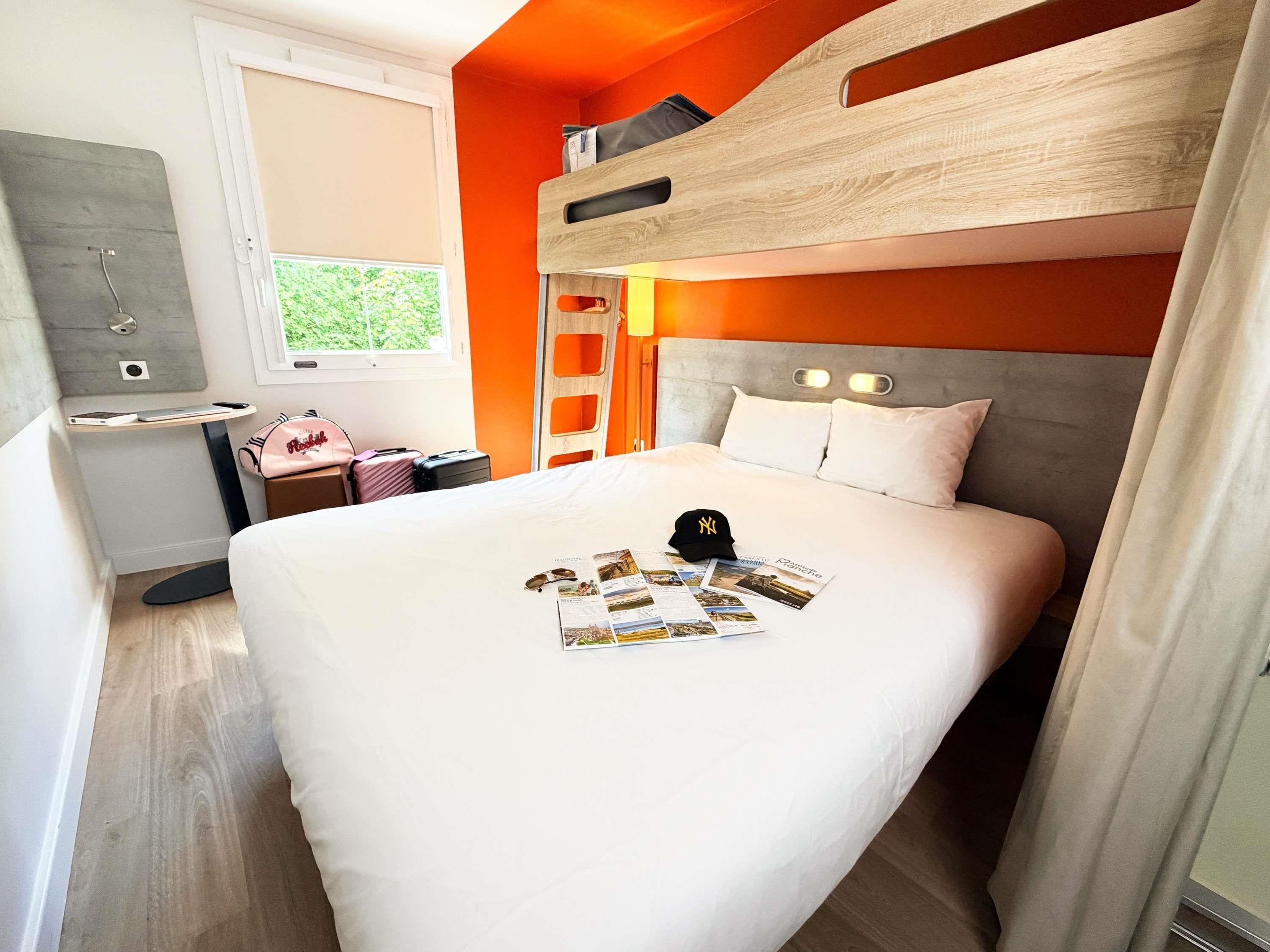 Et moderne hotelværelse med køjeseng, et sengebord, en kuffert og et vindue med udsigt til grønne områder.