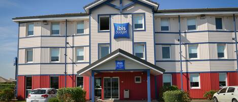 Un hotel de varios pisos con un cartel llamativo de 'ibis budget', una entrada cubierta y un estacionamiento con automóviles.